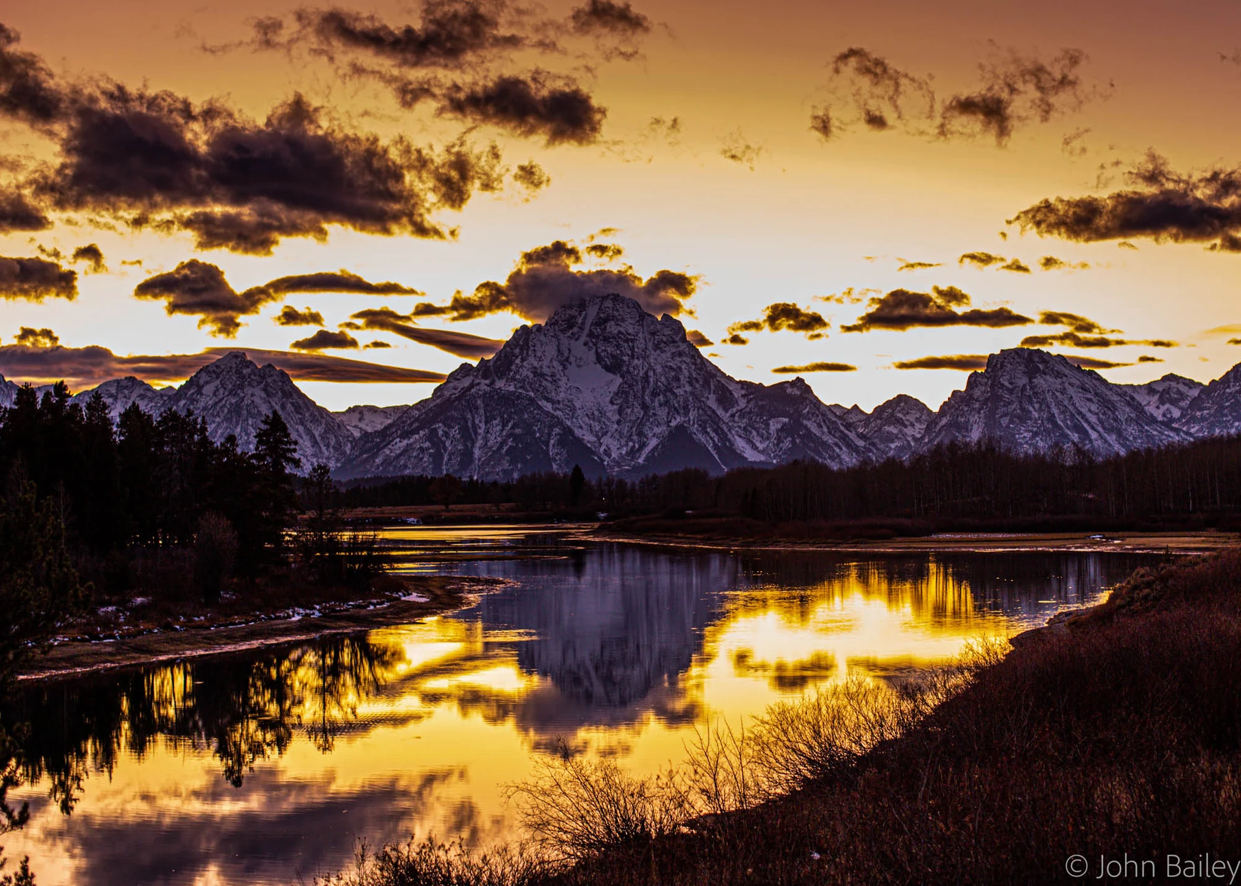 The Teton Range at sunset, jagged peaks glowing gold and reflected in the still water of the Snake River below