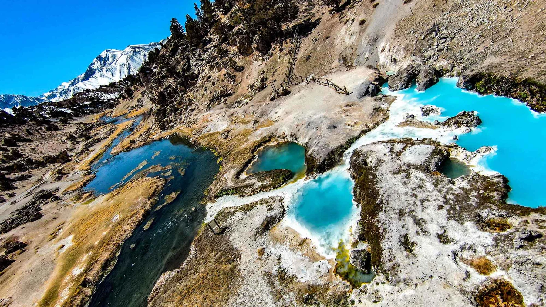 Aerial view of vivid turquoise glacial pools set in grey rock beneath a snow-capped ridge