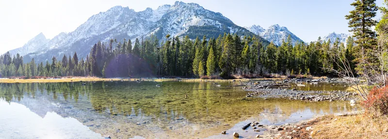 Panoramic view of a snow-dusted mountain range above a clear glacial lake and pine forest