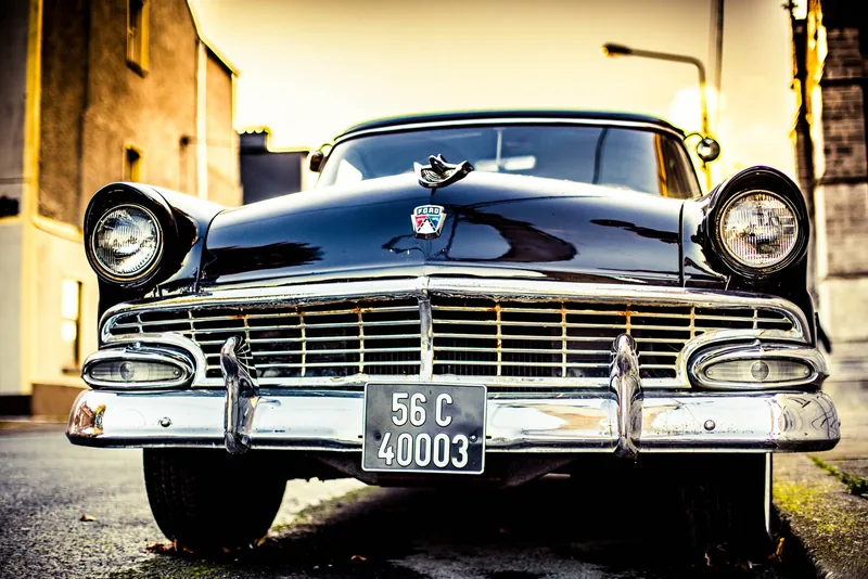 The chrome grille and hooded headlights of a 1956 Ford in evening light on a cobbled street