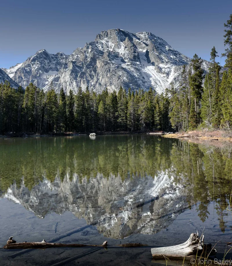 A granite peak and dense pine forest reflected in a perfectly still alpine lake