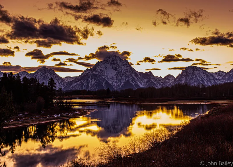 The Teton Range at sunset, jagged peaks glowing gold and reflected in the still water of the Snake River below