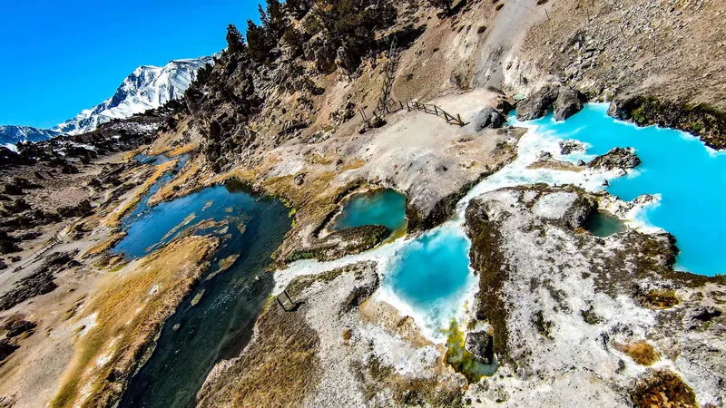 Aerial view of vivid turquoise glacial pools set in grey rock beneath a snow-capped ridge