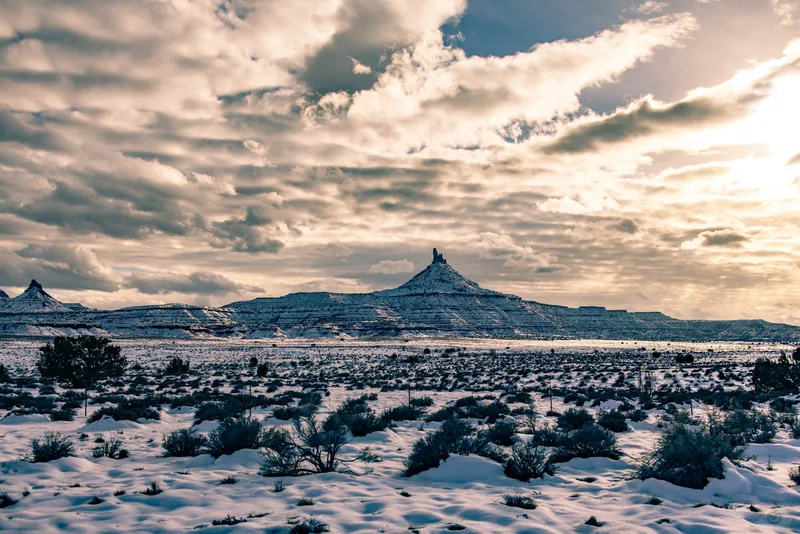 A lone volcanic butte rising from a snow-covered desert plain under dramatic cloud
